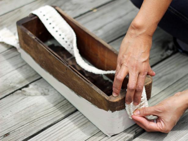 Step 4 in crafting a chic bath caddy for guests is preparing to fasten the fabric tape, cut to length, to each side of the painted and dry wooden box. Eventually the trim will be fastened to a doorknob with a nut and bolt.
