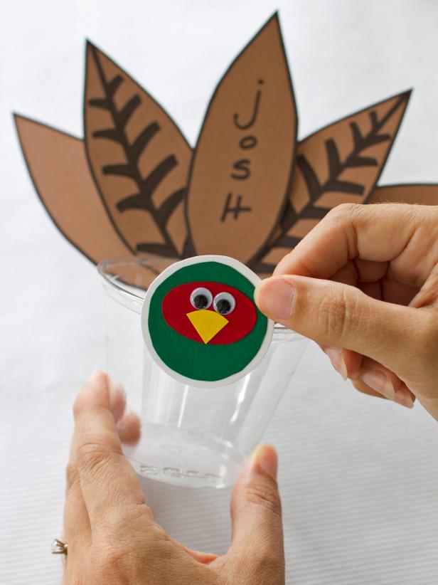 After you have secured the feathers on your Thanksgiving place cups, use hot glue to secure the turkey face to the front edge of the cup.