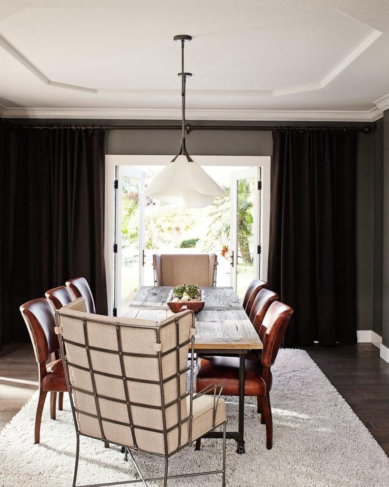 Two sets of French doors open this dining room to a lovely outdoor setting, while dark curtains provide drama and frame the view. A rustic-industrial table is surrounded by a mix of seating for eclectic charm.