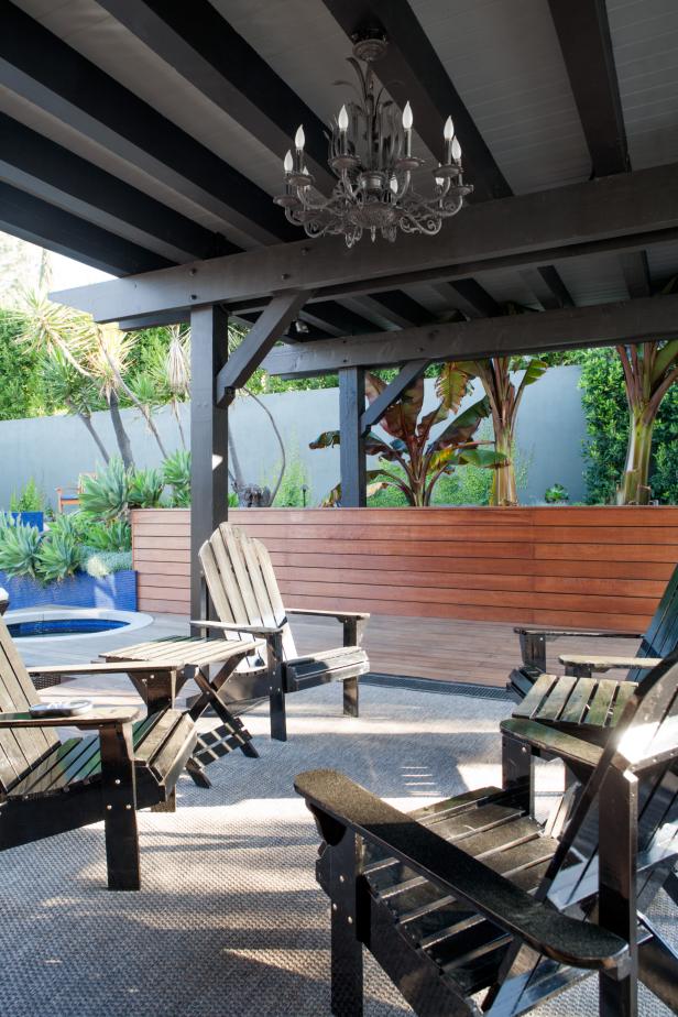 This spacious backyard features a small sitting area with black Adirondack chairs and end tables, and a gray area rug. A pergola painted black and gray and finished with a glamorous chandelier tops off the look of the outdoor space.