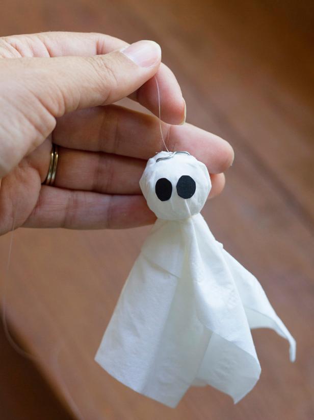 To create a spooky ghost to add to a Halloween-inspired terrarium, tear a tissue in half and layer the pieces to form a cross, then place a 1&quot; foam ball in the center. Gather the tissue around the foam ball and use fishing line to create the ghost's neck by tying a knot around the base of the foam ball. Cut out two black tag board ovals to act as eyes and attach them with glue. Shorten the prongs of a U-shaped floral pin with wire snips, and then push it into the top of the ghost's head. Knot one end of a piece of fishing line around the floral pin and attach the other end to the underside of the jar's lid with clear tape.