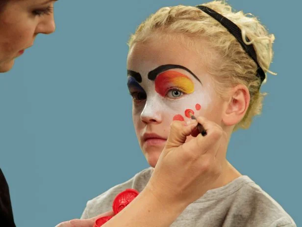 In order to give your child the perfect clown makeup for Halloween, use a makeup sponge and brush to apply white makeup to just the eyes and nose. Feather the makeup around the outside edges of the face so it fades softly. Use a makeup brush and blue face paint to draw a half circle over one eye following the brow line. Blend the blue and white. Do the same thing to half of the other eye, using red face paint. Next, add yellow face paint to the rest of the eyebrow and the upper lid. Blend slightly. Use a black eyeliner pencil to make a half circle above the child's natural eyebrow, then a line that follows the natural brow. Use a craft brush with red paint to make three small circles on one cheek. On the opposite cheek, draw a large circle with pink face paint. To make a clown mouth, first use red lip liner to line just outside the lips, then fill in. Extend the lines out from the edge of the natural lips and add little circles at the end. Outline lips with black liquid eyeliner. As a final accent, draw some lines just below the lower eyelid. Naturally curly hair (or a wig) is ready to clown around and just needs a nose. Oversized clothes or a clown costume finishes this big-top look.
