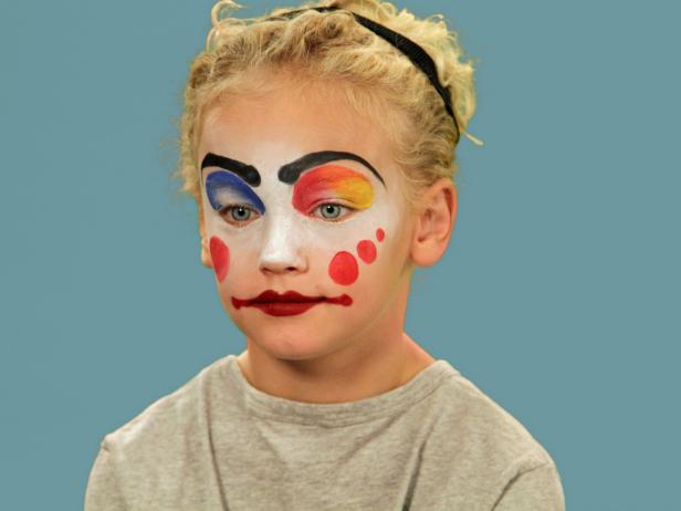 In order to give your child the perfect clown makeup for Halloween, use a makeup sponge and brush to apply white makeup to just the eyes and nose. Feather the makeup around the outside edges of the face so it fades softly. Use a makeup brush and blue face paint to draw a half circle over one eye following the brow line. Blend the blue and white. Do the same thing to half of the other eye, using red face paint. Next, add yellow face paint to the rest of the eyebrow and the upper lid. Blend slightly. Use a black eyeliner pencil to make a half circle above the child's natural eyebrow, then a line that follows the natural brow. Use a craft brush with red paint to make three small circles on one cheek. On the opposite cheek, draw a large circle with pink face paint. To make a clown mouth, first use red lip liner to line just outside the lips, then fill in. Extend the lines out from the edge of the natural lips and add little circles at the end. Outline lips with black liquid eyeliner. As a final accent, draw some lines just below the lower eyelid. Naturally curly hair (or a wig) is ready to clown around and just needs a nose. Oversized clothes or a clown costume finishes this big-top look.