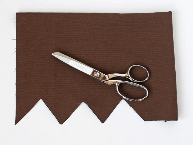 Lay out a piece of brown fabric and fold in half so that when hung next to the child, it will hang to the child’s mid thigh.Using scissors, cut along the edge opposite from the folded edge in a zig-zag pattern. Cut through both layers of fabric.