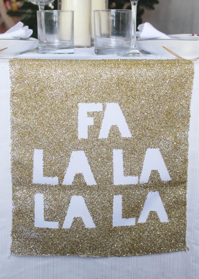 Do you like glitter? Then this is for you! No holiday decor is complete without the addition of something glittery. Youâ  ll need a white table runner, letter stickers, spray adhesive, and gold glitter. Spell out your saying, here I did Fa La La La La, with the sticky letters onto the white runner. Spray all around the letters with spray adhesive. Pour on your glitter. Youâ  ll want to push the glitter into the adhesive a little bit. Shake off the excess glitter.