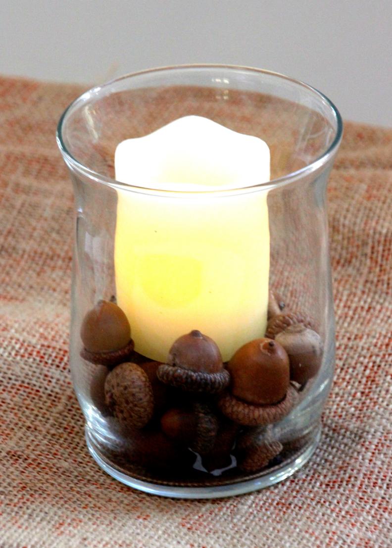 A glass votive jar is filled with acorns and topped with a candle.