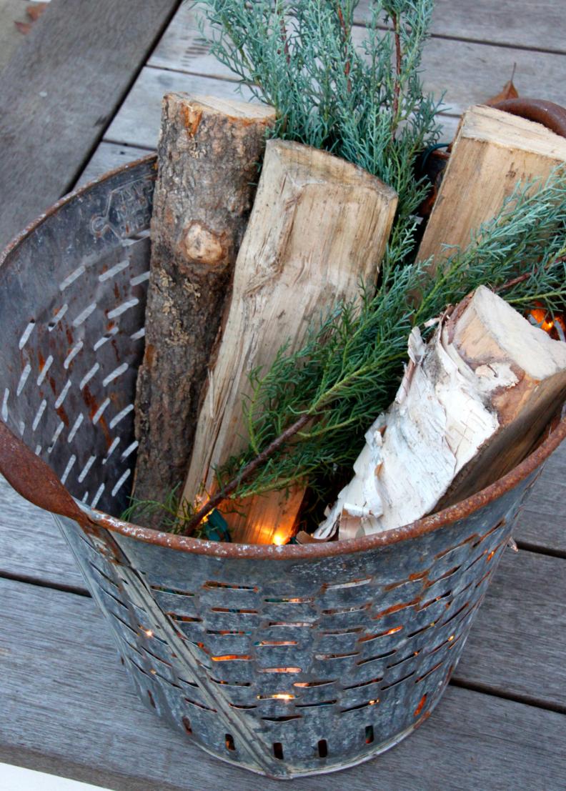 For a pretty front door porch display fill a stainless steel bucket, or an olive bucket in this case, with greenery, split logs and lights.
