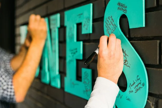 Press each letter up against the wall, ensuring even spacing. When guests arrive, have them use the black permanent markers to write messages and sign their names.