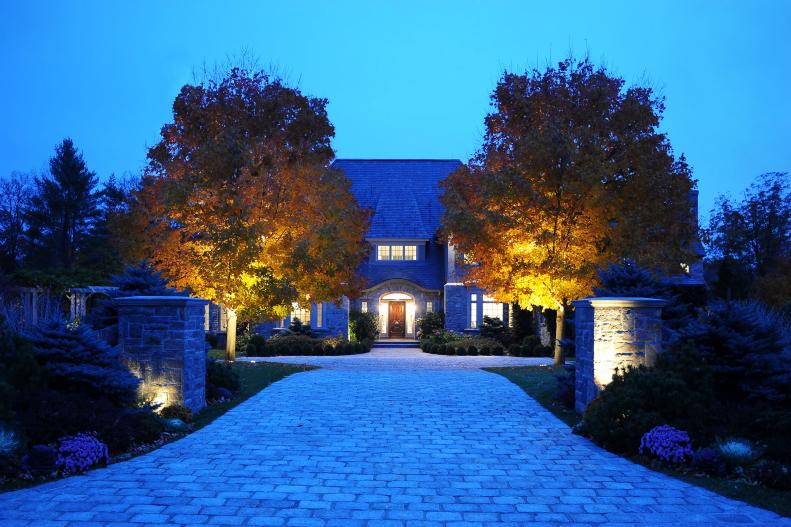 Stone Posts and Trees Flank Paver Driveway Leading to Gray Home
