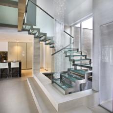 Modern Staircase With Glass Steps Wrapped Around Artistic Shower-Like Feature Falling Into Indoor Pond 