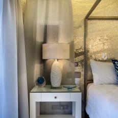 Modern Bedside Table With Texture Lamp in Front of Sheer Window Curtain and Sparkling Silver Accent Wall 