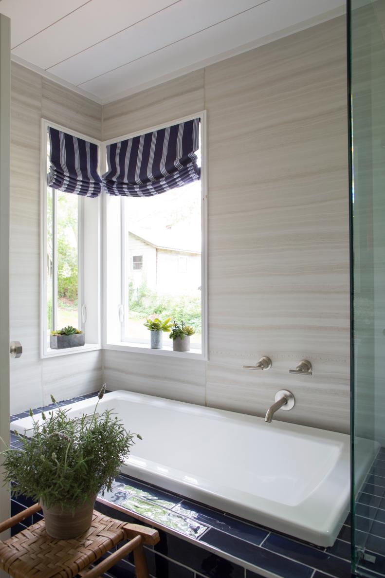 Navy porcelain tile engulfs this crisp white soaking tub, while a corner of windows introduces relaxing scenery. Beside the tub is a locally-sourced vintage tool to set lavender or other bath time amenities. 