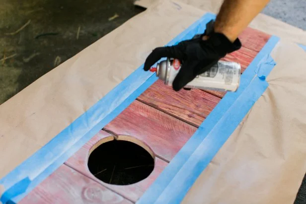 Using a palm sander and a fine grit paper, sand the rough spots down so that the bags will slide a bit when they hit the boards (Image 14). Dust off boards after sanding. To add vintage color, dip the tip of a dry brush into latex paint, barely getting paint on the bristles. Using this dry brush method, lightly sweep color onto the boards (Image 15). To add a white stripe, protect the sides using craft paper secured with painters tape. Lightly sweep white spray paint down the vertical center for an aged look (Image 16). Once dry, remove painters tape and craft paper, then …GAME ON!