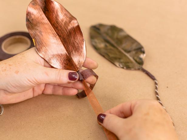 Use a paper towel to gently remove some of the preservative from the magnolia leaves. Then, using acrylic craft paints in metallic colors, lightly paint leaves in an imperfect way to give them a rustic look. After the leaves have fully dried, fasten a 9-12 inch length of sheer ribbon or yarn to the stem of each leaf, securing with brown floral tape. Add a dab of hot glue, as well for extra strength. Set leaves aside when done.