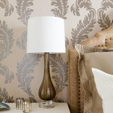 Off-white End Table with Blown-glass Lamp near Upholstered Headboard and Metallic-printed Wallpaper