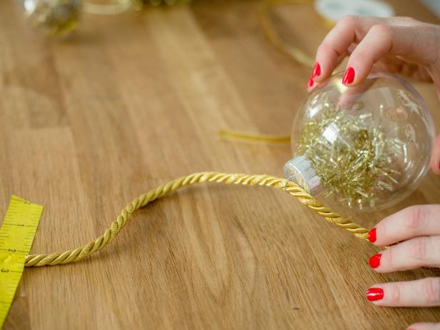 Once you have the ornaments prepped you can begin tying them onto the gold cord to create a garland. Tape one end of the cord to the table and slide ornaments on from the other direction toward the taped end. Secure the first ornament with a small piece of ribbon to keep it from sliding around. Do this every so often as you add more ornaments to the garland , then tie the very last ornament into place.