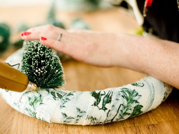 Remove the bases of the bottle brush trees and bushes so that the wire is exposed. Insert wire into the foam wreath, then bend so that the tree is flat against wreath, securing with hot glue as needed. Repeat with other trees and bushes until you get a landscape effect on either side of the wreath.