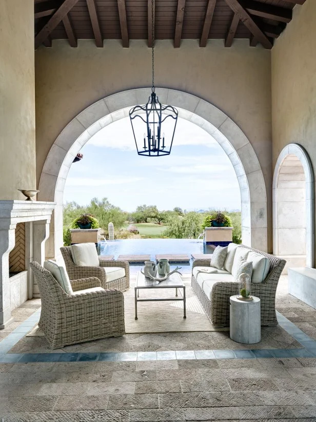Neutral wicker furniture is combined with a glass coffee table and elegant lantern chandelier for a comfortable outdoor seating area. Exposed wood ceiling, stucco walls and textured neutral brick patio evoke a southwestern feel, overlooking the beautiful pool area.