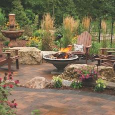 Paver Patio With Stone Fire Pit, Brown Adirondack Chairs and Mulched Surrounding Garden 