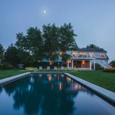 A Long Pool Reflects The Tree Line Over Beautiful White Home Exterior With Spacious Yard and Poolside Seating