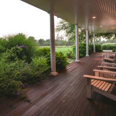 Long Covered Wood Porch With Tan Adirondack Chairs, White Columns and Thick Shrubbery 