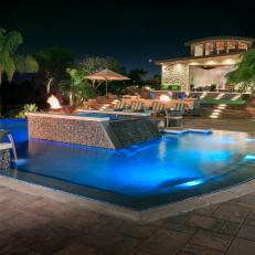 Nighttime View of Luxurious Tropical Swimming Pool With Waterfall Embellishments and Stone Tile Patio
