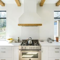 Bright White Cabinetry and Range Hood Surrounding Stainless Steel Stove Under Natural Wood Exposed Beams 