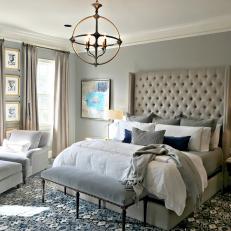 Large Beige Tufted Headboard Over Layered Pillows in Traditional Bedroom With Spherical Chandelier and Patterned Carpet