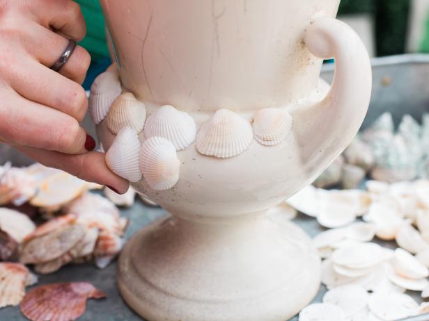 Start in one area of the urn and begin by adding a simple row of similar looking shells using the hot glue gun. You’re essentially creating bands up and down the urn- using one particular type of shell. Place a bead of hot glue onto the perimeter of the shell then push it into place on the urn. It helps to let the shape of your particular vessel and the shell dictate your design. At the base of the urn, leave a little space, so that the shell’s edge doesn’t chip off or scratch the surface of the table it’s sitting on and remember to rotate the shells in different directions, as well for visual interest and better coverage. We used shells in a variety of shapes and colors.