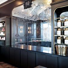 Distressed Mirror With Name Emblem and Brass Trimmed Shelves Over Black Cabinetry at Macaron Bakery Counter 