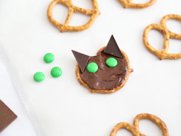 Arrange two triangles on the top portion of the pretzel for ears. Press them gently into the melted chocolate. Position the green candy-coated chocolates just under the ears for the eyes.