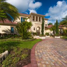 Brick Driveway Leading to Tropical Home Exterior With Palm Trees, Arched Entranceway and Balcony Columns 