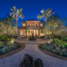 Grand Home Back Yard Exterior With Columned Balcony, Warm Lighting and Circular Stone Patio Framed By Tropical Trees 
