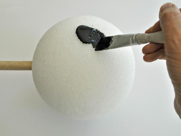 Align wooden dowel onto foam ball and trace around dowel with pencil. Use knife to carve out circular shape 1 inch deep. Hot glue end of dowel and place into hole of foam ball. Add additional glue to fill in around edges of hole. Repeat steps on other foam ball. Tip: Use coolest setting on hot glue gun to void melting foam. Paint barbell with black acrylic paint. Tip: Due to the porous nature of the foam ball, pour paint onto ball, then gently brush it around to coat all textured areas. Do not attempt to use spray paint; spray paint melts foam.