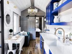 This tiny house is big on style! We enter into the kitchen and are immediately struck by the simple yet bold palette of cobalt blue, crisp white, and gray. Its contemporary vibe is grounded by the use of classic rustic elements. The brass details (current design trend), chunky shelves, cantilevered tables and the tufted sofa help to create an undeniably elevated look in this tiny home. I especially admire the restraint practiced by matching the white sink to the white counter to the white walls. It seems simple but it creates visual space, which is always valuable in a tiny home.  