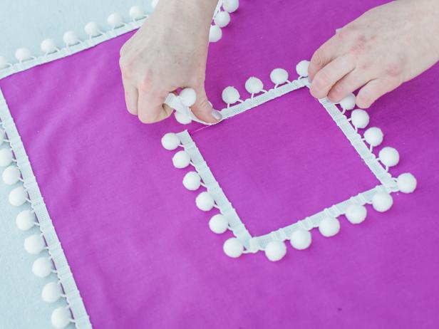 Step 11- Add Pom-Pom TrimCut trim to size for each “run” or side of a square, then press into place atop the fabric glue. Hold the trim in place allowing the glue to set. Start with the perimeter of the pillow, then create the innermost square of pom-pom trim.