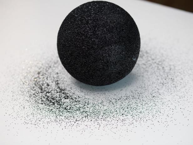 Set the medium or large Styrofoam ball on top of a sturdy surface. Use craft paper or a painters drop cloth underneath the Styrofoam balls before painting to protect your surface.  Completely cover the Styrofoam ball with the black spray paint.  Immediately after spray painting the Styrofoam ball while it is still wet pour on the black glitter, let dry.