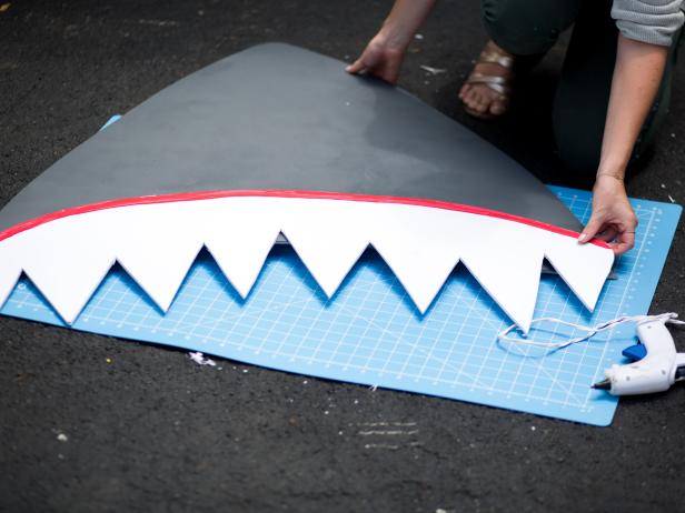 Drape a red sheet over the open trunk to help create a backdrop for the shark’s mouth.  With the hot glue gun attach the top teeth to the snout and press firmly.  Attach the two dowel rods to the snout with painters tape. This will support the snout and hold it up out of the trunk. Then, set the bottom teeth in front of the dowel rods and secure them with painters tape.