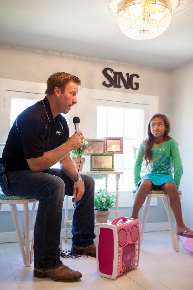 Host Chip Gaines shows Charmaine and Chuck’s daughter Charlie how her new karaoke machine works by singing Charlie a love song.  Charlie is not impressed with Chip’s singing, but she is excited her new room on the third floor, complete with a stage, a reading nook, a desk for doing homework, and a table and chairs.  Hosts Chip and Joanna also made a custom dollhouse for Charlie, as seen on HGTV’s Fixer Upper. (action-reveal)