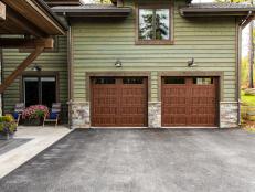 A durable asphalt driveway leads to the insulated panel-style garage doors made from durable and low-maintenance steel with window inserts and a walnut color finish at the front of the garage, with farmhouse-style gooseneck light sconces above that illuminate the area. 