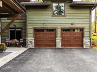 A durable asphalt driveway leads to the insulated panel-style garage doors made from durable and low-maintenance steel with window inserts and a walnut color finish at the front of the garage, with farmhouse-style gooseneck light sconces above that illuminate the area. 