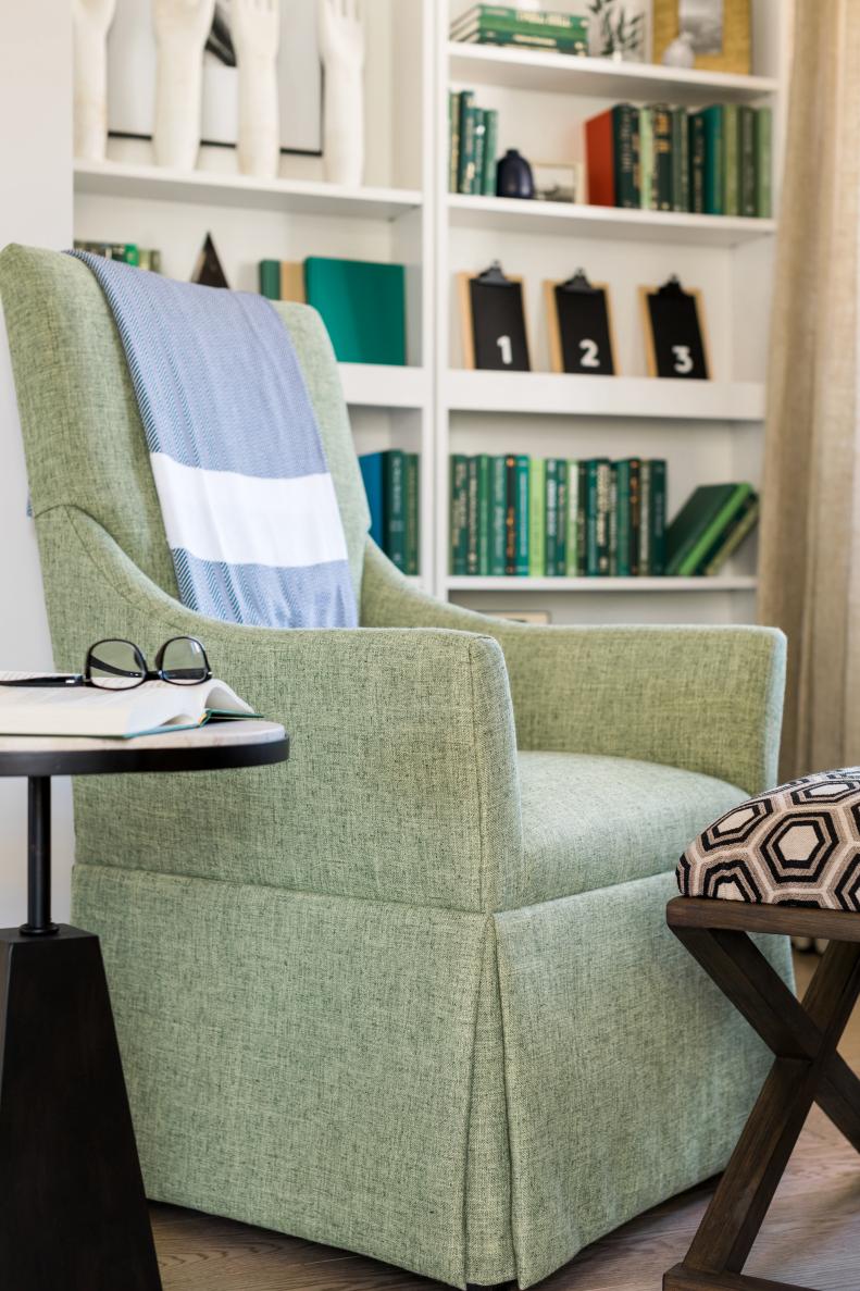 A modern upholstered side chair with a skirted base is paired with an upholstered stool to create comfortable seating in the reading area. A side table with a travertine stone top offers a spot for resting a book or drink. 