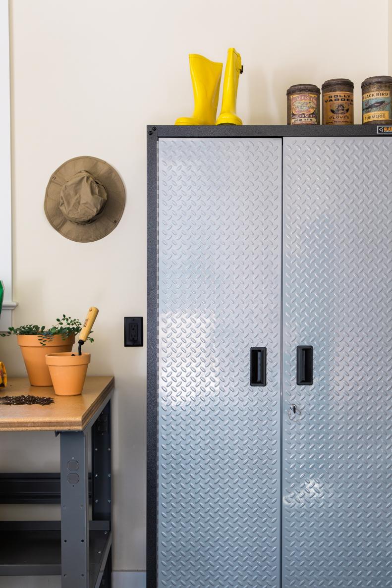 The heavy-duty garage cabinets feature a hammered granite finish with silver tread. On top of the cabinets sit various garden items, including clay pots in assorted sizes and a pair of yellow rain boots for wet days out in the yard. 