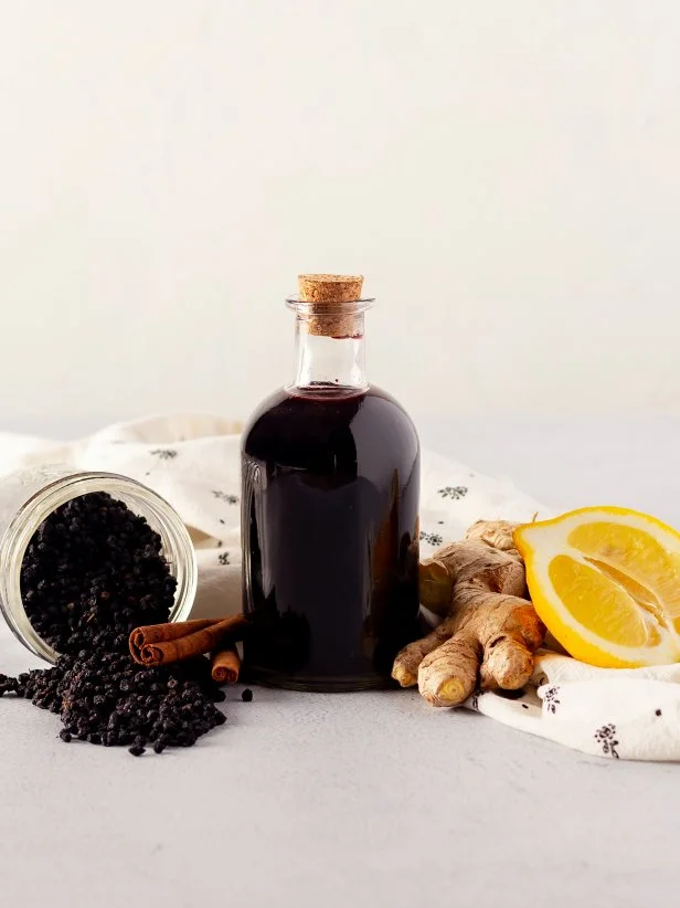 A bottle of elderberry syrup with dried elderberries, ginger, lemon and cinnamon sticks. 