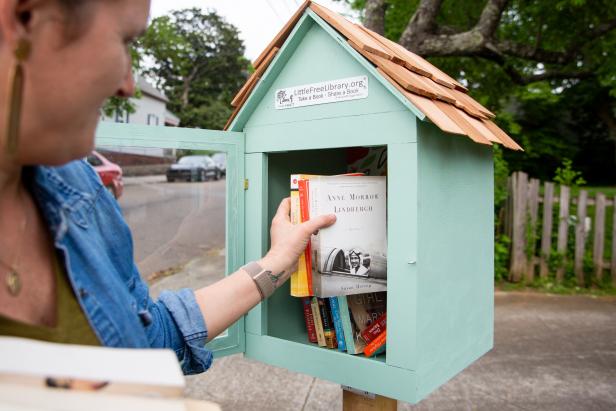A woman is sliding a handful of books into the little free library that she built. The library is light blue and features a shingled roof with a glass door that is ajar in the photo.