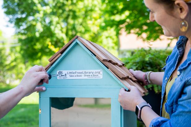 Two people screw the official sign on the DIY &quot;Little Free Library&quot; that they created. The library is light blue and features a glass door and a shingled roof.