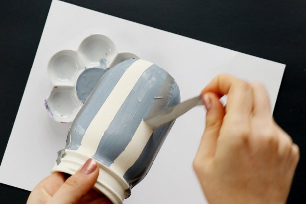 Mask off vertical stripes on the jar using washi tape. Mix white, blue, and black paint to get a grayish-blue color. Paint between the tape and remove the tape to reveal vertical stripes. Let dry completely.