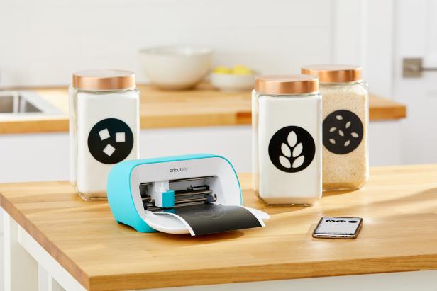 Cricut Joy sits on a wooden table and cuts black vinyl labels for canisters. 