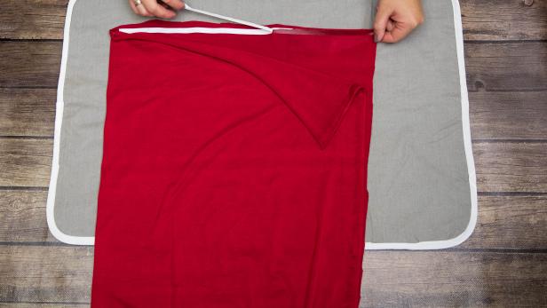 The paper backing is being pulled from this iron-on hem tape to attach this red jersey fabric to itself. The fabric is used to create a Little Red Riding Hood Halloween costume.