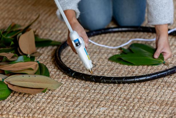 This hot glue is applied to a hula hoop that has been painted black as part of an oversized Christmas wreath project. The hot glue will hold bundles of foraged magnolia leaves to the hula hoop.